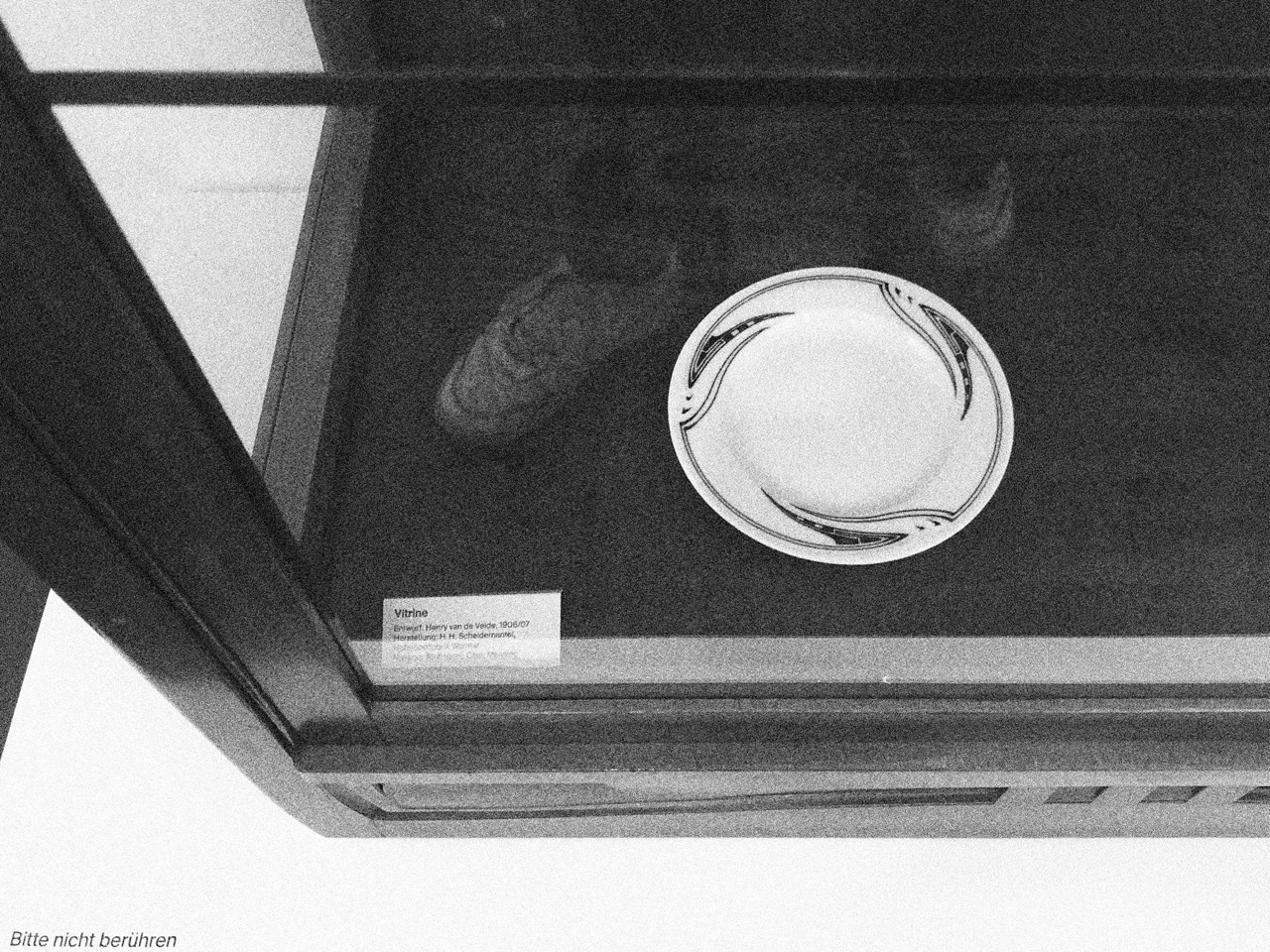 Schwarz-Weiß-Fotografie eines Ausschnitts einer Vitrine mit spiegelnder Glasscheibe, darin ein Teller mit Dekor und Objektbeschriftung: Entwurf: Henry van de Velde 1906/07, Herstellung: Der restliche Text ist wegen der Spiegelung nicht lesbar.