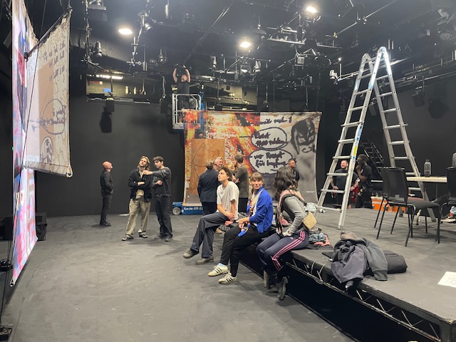 Mehrere Personen in einem großen Studio mit Bühnenbeleuchtung, einige sitzen auf einer erhöhten Bühne, andere stehen davor, im Hintergrund eine Leiter und große bedruckte Stoffbanner