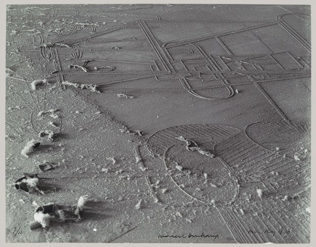 Schwarzweißfotografie einer staubbedeckten Glasplatte aus der Vogelperspektive. Die Oberfläche ist von einer feinen, ungleichmäßigen Staubschicht überzogen, die wie eine karge, mondähnliche Landschaft wirkt. In den Staub zeichnen sich geometrische Linien und Formen ab – gerade Strecken, Kreise und ovale Umrisse –, die technisch und konstruiert erscheinen. Die Linien wirken wie freigelegte Konturen unter der Staubschicht. Im linken unteren Bereich liegen mehrere kleine, weiche Staubballen oder Flocken, die sich von der flachen Fläche plastisch abheben. Rechts unten ist ein großer ovaler Bereich mit parallelen, gerillten Strukturen sichtbar, die an Felder oder technische Schraffuren erinnern. Am unteren Bildrand befindet sich eine schmale weiße Rahmung mit handschriftlichen Notizen und einer Signatur. 