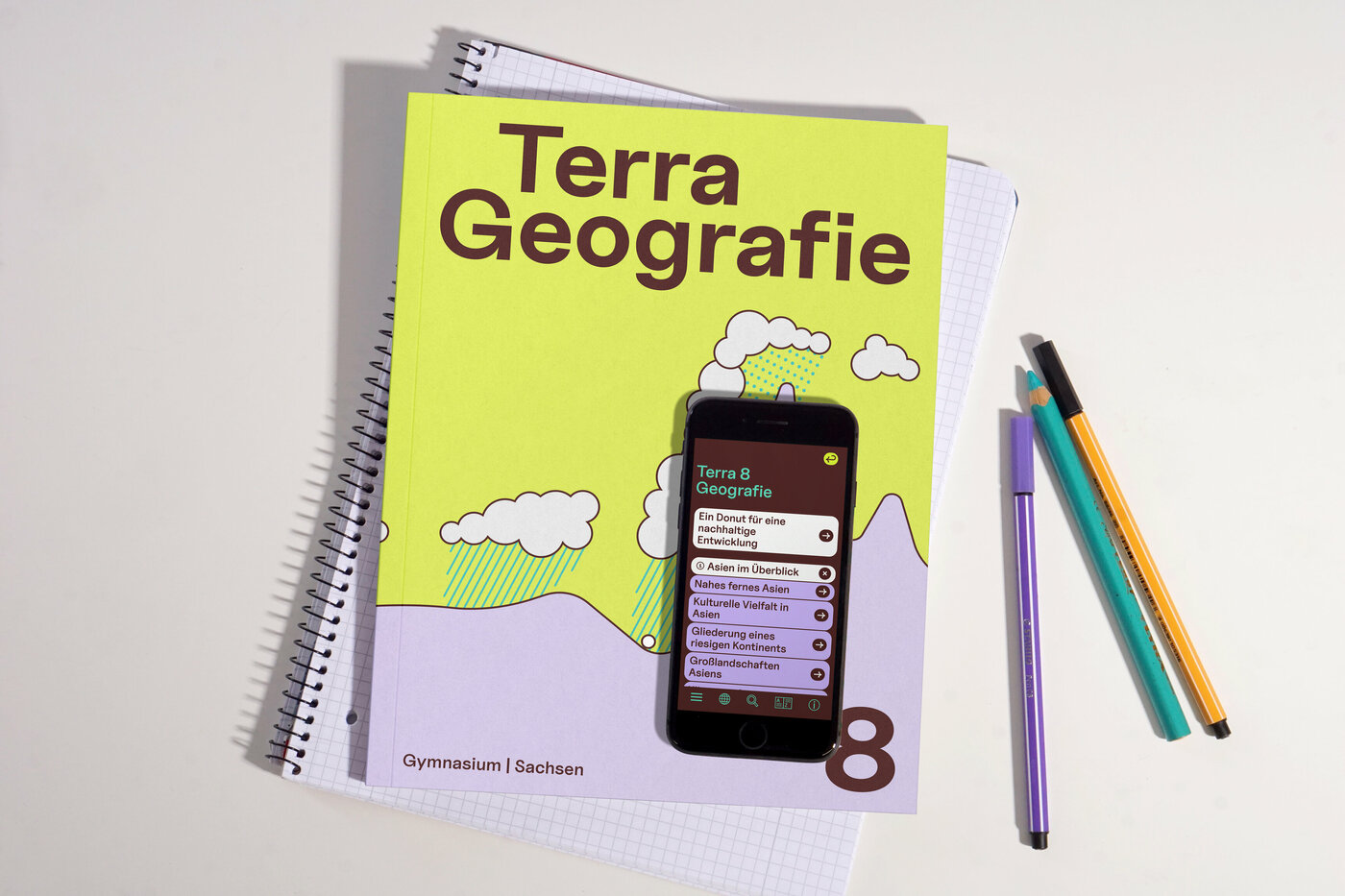 Das Bild zeigt das neu gestaltete Schulbuch „Terra Geografie 8“ für Gymnasien in Sachsen. Das limonengelbe Cover des Buches bildet neben dem Titel eine illustrative Grafik mit fliederfarbenen Bergen, hellgrauen Wolken und grünen Regentropfen ab. Auf dem Buch liegt ein Smartphone, auf dem das Menü der digitalen App zum Schulbuch geöffnet ist. Im Menü sind die verschiedenen Kapitel- und Themenüberschriften zu sehen. Der Hintergrund-Screen des Menüs ist in einem dunklen Braun gehalten und die Überschriften befinden sich in limonengelben und fliederfarbenen Kästen mit Navigationselementen wie Plus-Symbolen oder Pfeilen. Das Schulbuch liegt auf einem Notizblock, daneben sind drei Stifte platziert.