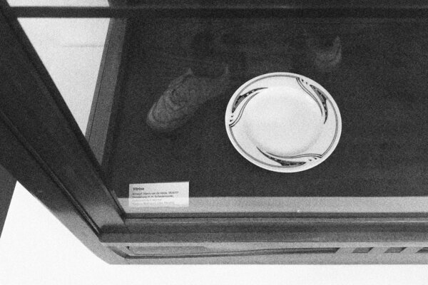 Schwarz-Weiß-Fotografie eines Ausschnitts einer Vitrine mit spiegelnder Glasscheibe, darin ein Teller mit Dekor und Objektbeschriftung: Entwurf: Henry van de Velde 1906/07, Herstellung: Der restliche Text ist wegen der Spiegelung nicht lesbar.