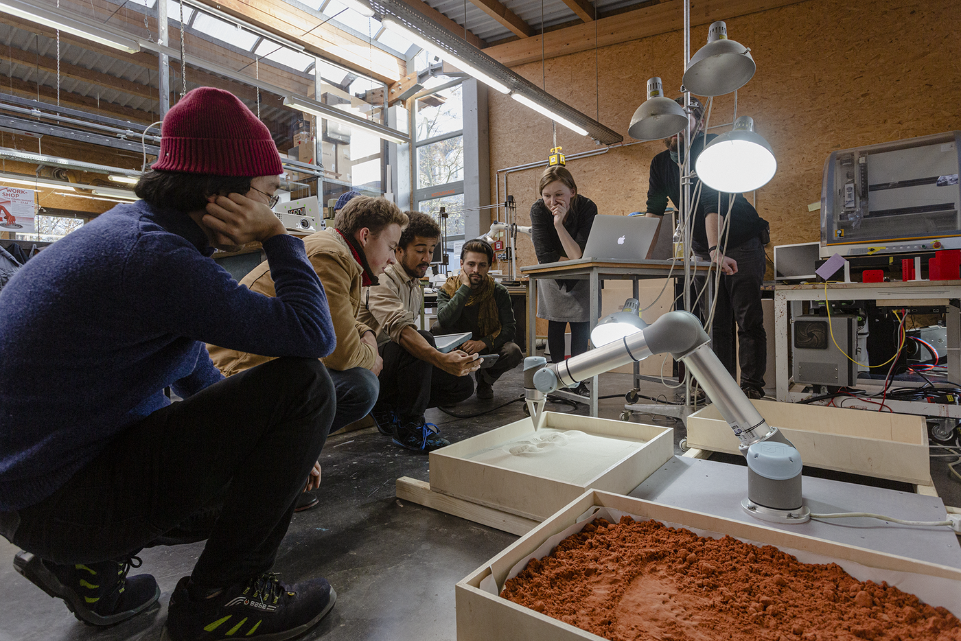 eine Kurs von Studierenden beobachtet einen Roboterarm, der mit montierter Metallspitze Muster in ein Sandbecken zeichnet
