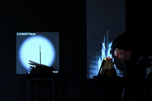 In zwei Installationen werden analoge Objekte mitteles einer Projektion an der Wand augmentiert. Ein Mensch interagiert mit einer der Installationen in dem er in einen Holzhaufen pustet. In der Projektion wird ein Feuer angefacht.  