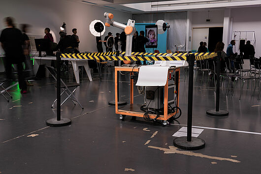 Zu sehen ist ein Roboterarm, umgeben von einem gelb-schwarzen Absperrband.  Im Hintergrund sind weitere Installationen zu sehen. Viele Menschen stehen verteilt im Raum und betrachten die Installationen.