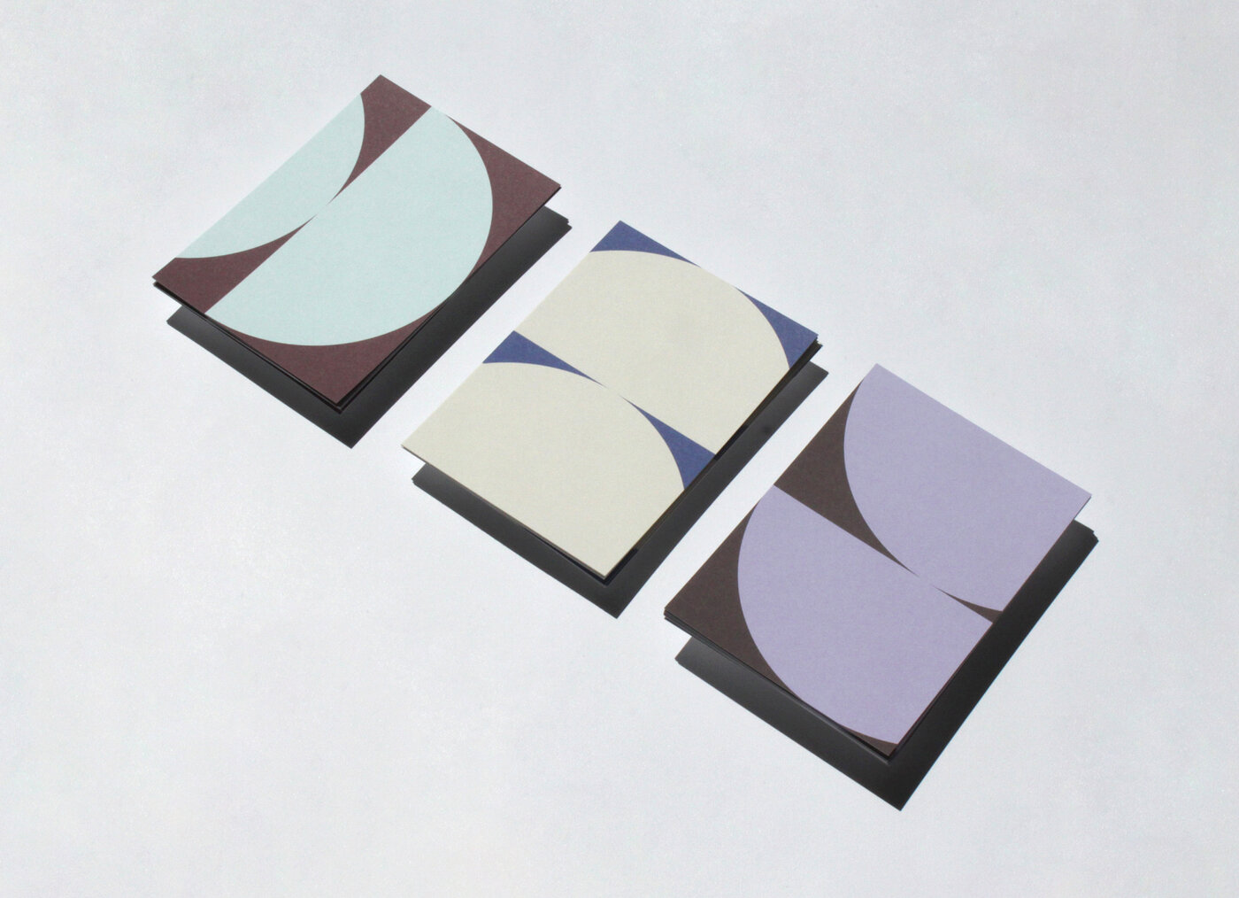 Das Bild zeigt eine Fotografie von drei nebeneinanderliegenden Postkarten. Auf den Postkarten sind zwei doppelte Halbkreis-Formen zu erkennen. Die linke Postkarte hat einen dunkelroten Hintergrund und eine hellblaue Form. Die zweite Karte hat einen dunkelblauen Hintergrund und eine hellgelbe Form. Die dritte Karte hat einen dunkelbraunen Hintergrund und eine fliederfarbene Form.