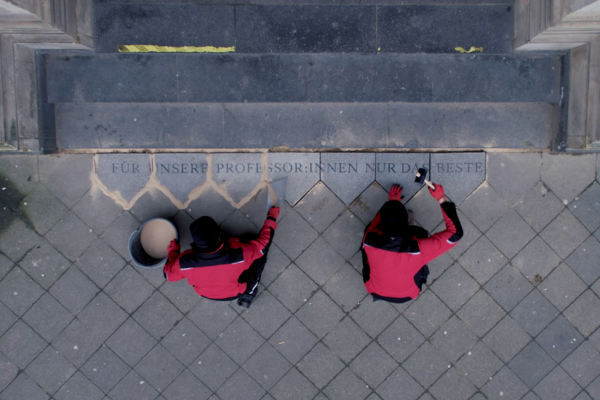 Die Fotografie zeigt eine Draufsicht auf den Eingangsbereich eines Gebäudes mit breiter Steintreppe. Vor der untersten Stufe sitzen zwei Personen auf dem Boden, beide tragen schwarze Hosen, schwarze Mützen und rote Jacken mit schwarzen Ärmeln. Sie arbeiten an Bodenplatten, die direkt an die Treppe angrenzen. Eine der Personen hält einen runden Eimer und eine Schaufel vor sich, die andere einen Gummihammer. In die Steinplatten ist ein Schriftzug eingraviert, der sich über mehrere Platten erstreckt. Er lautet: „FÜR UNSERE PROFESSOR:INNEN NUR DAS BESTE“. Einige Fugen zwischen den Steinen sind frisch verfugt und heben sich hell vom grauen Pflaster ab.