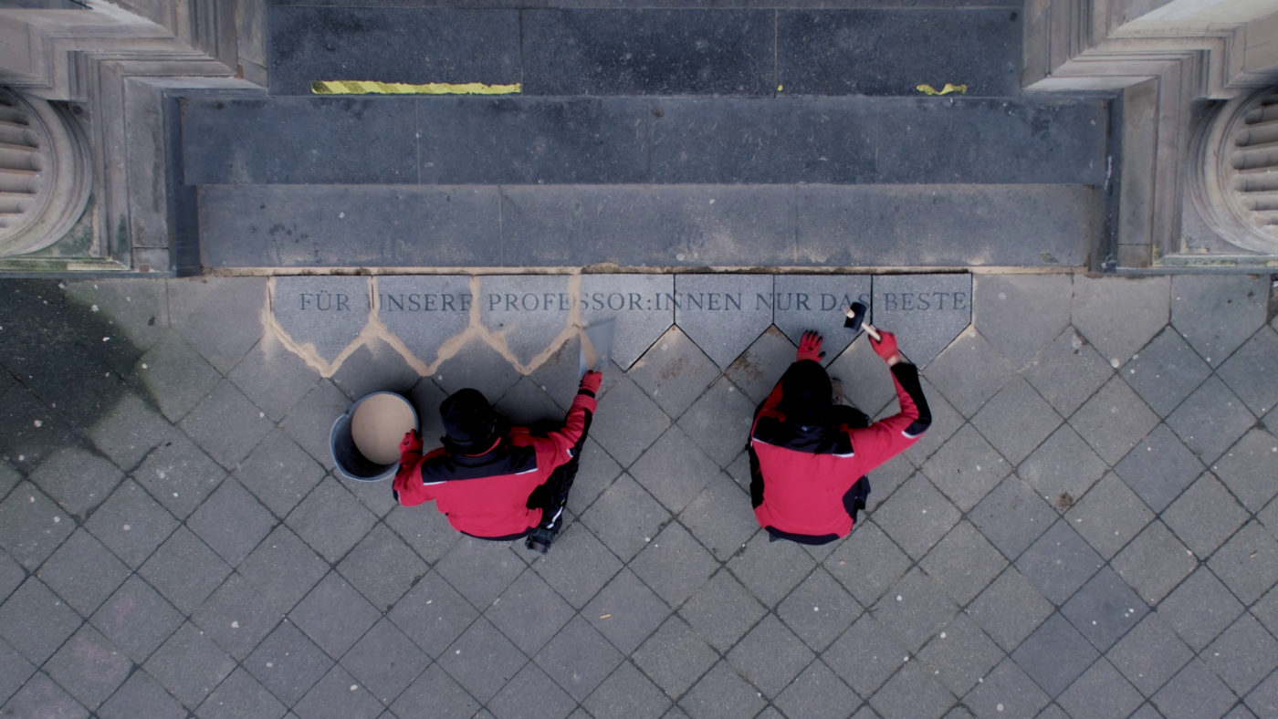 Die Fotografie zeigt eine Draufsicht auf den Eingangsbereich eines Gebäudes mit breiter Steintreppe. Vor der untersten Stufe sitzen zwei Personen auf dem Boden, beide tragen schwarze Hosen, schwarze Mützen und rote Jacken mit schwarzen Ärmeln. Sie arbeiten an Bodenplatten, die direkt an die Treppe angrenzen. Eine der Personen hält einen runden Eimer und eine Schaufel vor sich, die andere einen Gummihammer. In die Steinplatten ist ein Schriftzug eingraviert, der sich über mehrere Platten erstreckt. Er lautet: „FÜR UNSERE PROFESSOR:INNEN NUR DAS BESTE“. Einige Fugen zwischen den Steinen sind frisch verfugt und heben sich hell vom grauen Pflaster ab.