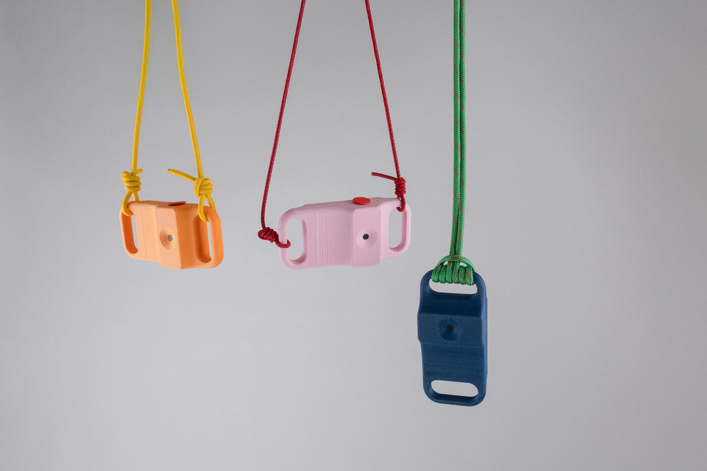 Drei kleine, 3D-gedruckte Video-Kameras in Orange, Rosa und Dunkelblau mit buntem Unhängeseil, hängen im Bild