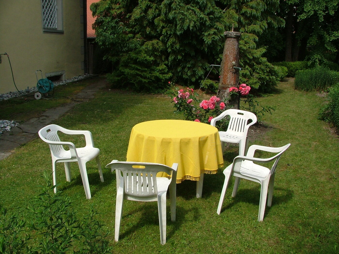 In einem Garten stehen vier Monobloc-Plastikstühle um einen mit einer gelben Tischdecke bedeckten Plastiktisch.