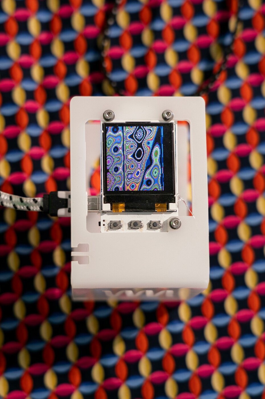 Dieses Bild zeigt die Installation Automaton von Tristan Schulze. Zu sehen ist ein kleiner Display, der ein Muster zeigt, vor dem Hintergrund eines Textils mit quadratischen Mustern.