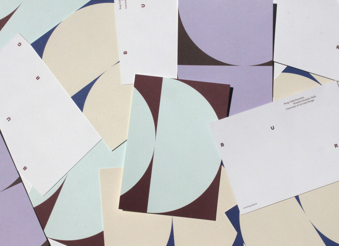 Das Bild zeigt eine Fotografie von wild durcheinanderliegenden Postkarten. Auf den Postkarten sind zwei doppelte Halbkreis-Formen zu erkennen. Eine Postkarte hat einen dunkelroten Hintergrund und eine hellblaue Form. Eine Karte hat einen dunkelblauen Hintergrund und eine hellgelbe Form. Eine Karte hat einen dunkelbraunen Hintergrund und eine fliederfarbene Form.