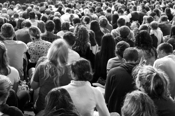 Fotografie in schwarz-weiß, auf dem Foto ist eine große Menschen an sitzenden Menschen von Hinten zu sehen, sodass man die Rücken, Köpfe und Arme der Personen sieht.
