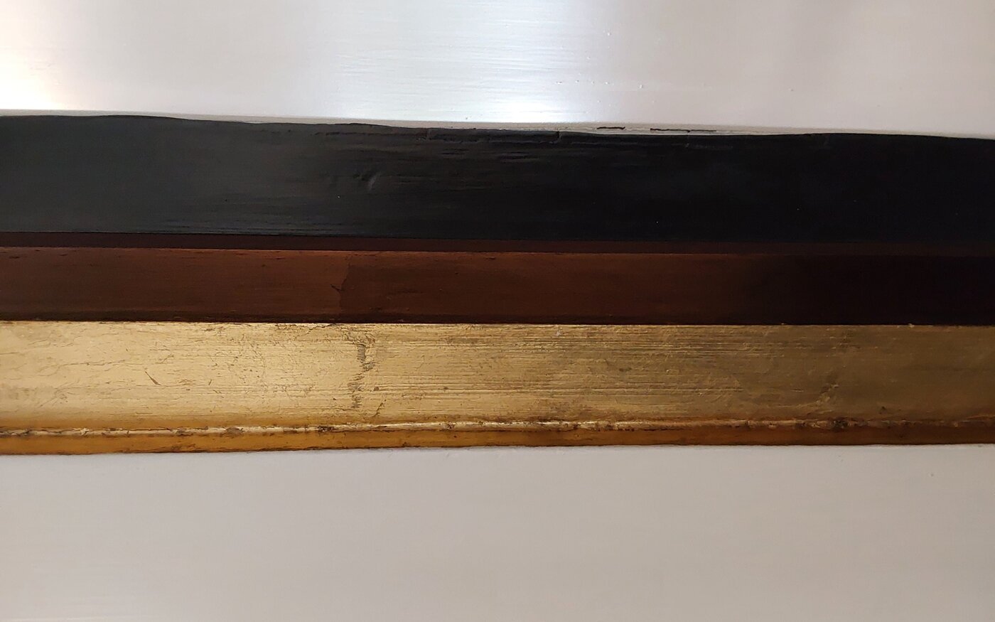 Das Foto "Gold, Braun, Schwarz" besteht aus drei senkrechten Streifen in Gold, Braun und Schwarz vor einem hellen Hintergrund. Die Farbe ist auf Holz gestrichen, dessen Struktur zu erkennen ist.