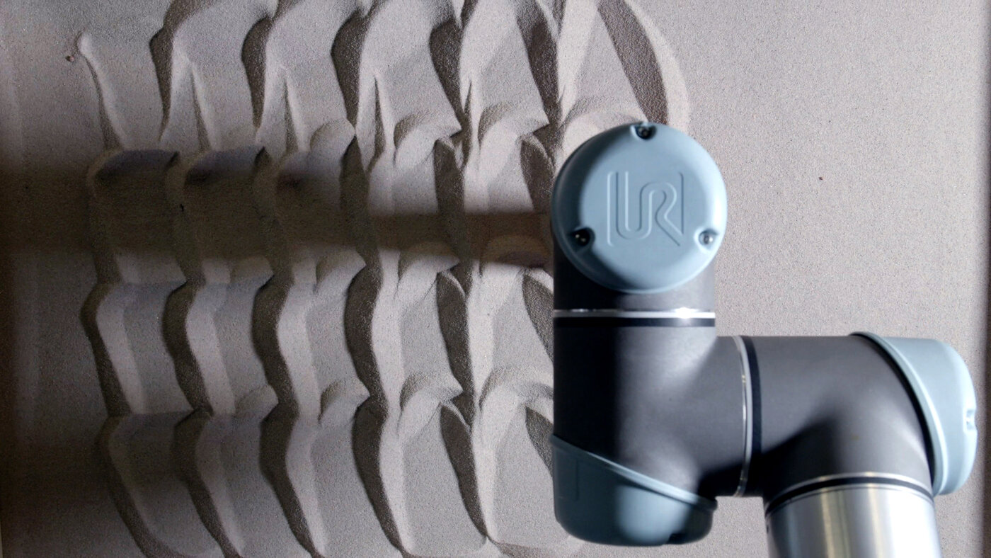 Nahaufnahme vom Roboterarm über einer Sandfläche. Der Roboter zeichnet eine parametrische Textur in den Sand - Foto BurgLabs