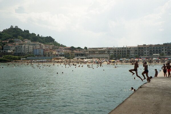 Weite Aufnahme einer belebten Badebucht an einem sonnigen Tag. Im Vordergrund rechts ragt ein betonierter Steg ins ruhige, grünlich schimmernde Meer. Zwei Jugendliche springen gerade vom Steg ins Wasser – ihre Körper sind in der Luft angewinkelt, die Arme erhoben, als hielten sie sich im Gleichgewicht. Neben ihnen stehen weitere Kinder oder Jugendliche am Rand und beobachten oder bereiten sich ebenfalls auf einen Sprung vor. Im Wasser verteilen sich zahlreiche Badende; viele Köpfe und Schultern ragen aus der Oberfläche. Im Hintergrund erstreckt sich ein breiter Sandstrand mit dicht an dicht stehenden Sonnenschirmen in hellen Farben. Dahinter stehen mehrstöckige Wohnhäuser mit Balkonen. Links erhebt sich ein grüner Hügel, auf dessen Spitze ein größeres Gebäude oder eine kleine Festung steht. Der Himmel ist leicht bewölkt, das Licht wirkt warm und sommerlich. Insgesamt vermittelt die Szene eine lebhafte, unbeschwerte Urlaubsatmosphäre.