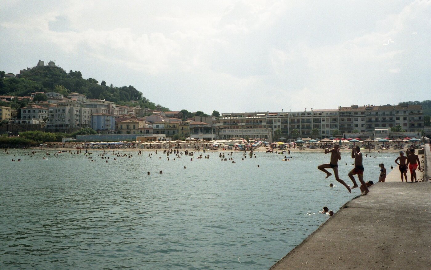 Weite Aufnahme einer belebten Badebucht an einem sonnigen Tag. Im Vordergrund rechts ragt ein betonierter Steg ins ruhige, grünlich schimmernde Meer. Zwei Jugendliche springen gerade vom Steg ins Wasser – ihre Körper sind in der Luft angewinkelt, die Arme erhoben, als hielten sie sich im Gleichgewicht. Neben ihnen stehen weitere Kinder oder Jugendliche am Rand und beobachten oder bereiten sich ebenfalls auf einen Sprung vor. Im Wasser verteilen sich zahlreiche Badende; viele Köpfe und Schultern ragen aus der Oberfläche. Im Hintergrund erstreckt sich ein breiter Sandstrand mit dicht an dicht stehenden Sonnenschirmen in hellen Farben. Dahinter stehen mehrstöckige Wohnhäuser mit Balkonen. Links erhebt sich ein grüner Hügel, auf dessen Spitze ein größeres Gebäude oder eine kleine Festung steht. Der Himmel ist leicht bewölkt, das Licht wirkt warm und sommerlich. Insgesamt vermittelt die Szene eine lebhafte, unbeschwerte Urlaubsatmosphäre.