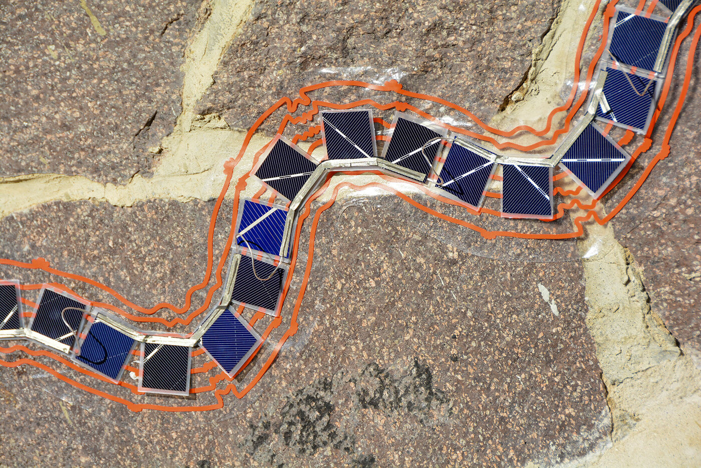 eine Reihe kleiner Solarpanele vor einer Steinmauer, in Wellenform eingeschweißt und mit grafischen, orangenen Linien versehen