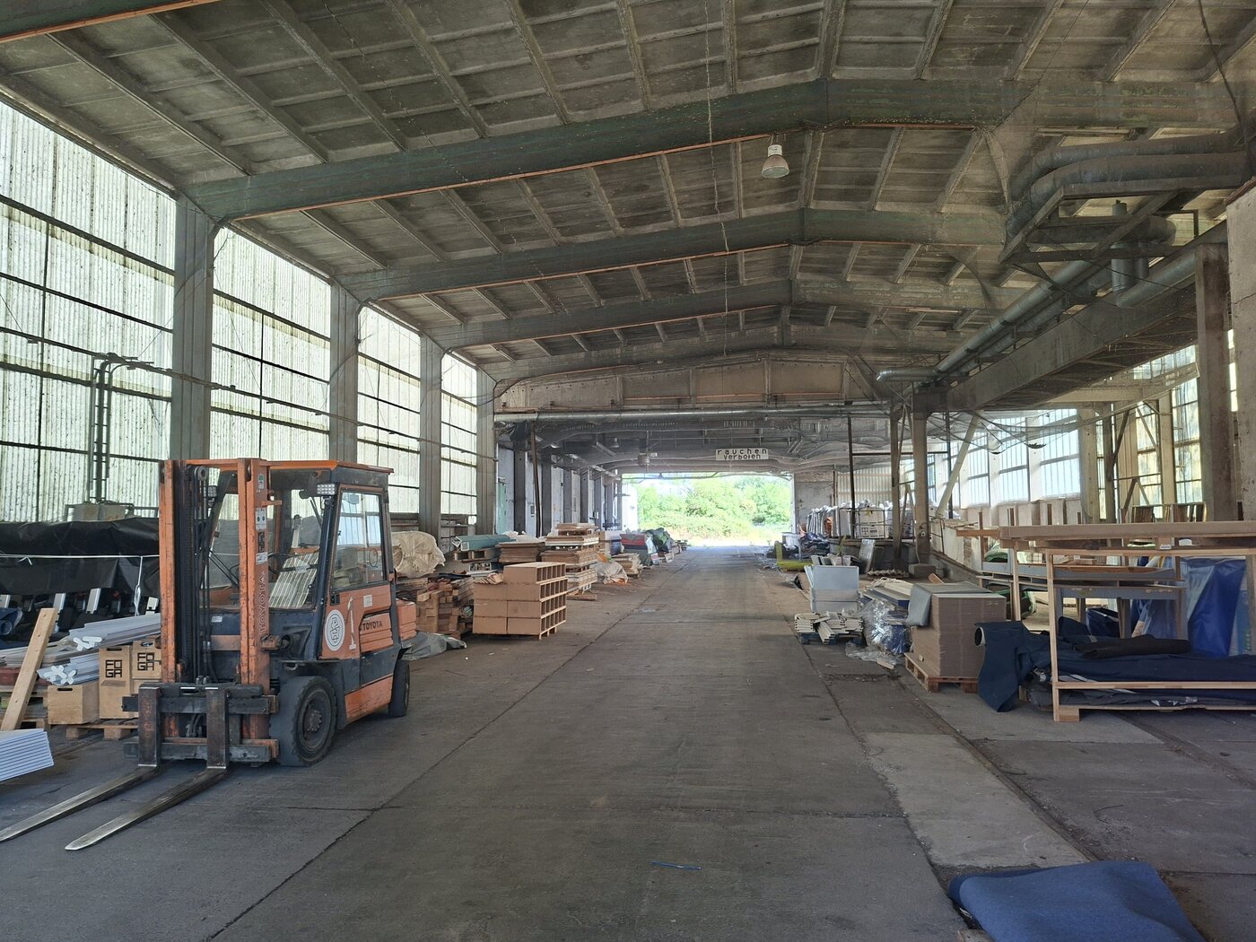 Blick in ein Materiallager einer Industriehalle. Links im Vordergrund steht ein orangefarbener Gabelstapler. Entlang beider lichtdurchlässigen Hallenseiten lagern unterschiedliche Materialien, Bauteile, Paletten und textile Elemente in offenen Regalen oder gestapelt auf dem Boden. Die Mitte der Halle bleibt als breite Fahr- und Bewegungsfläche frei und führt den Blick zu einer geöffneten Hallenseite im Hintergrund.