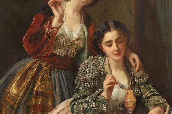 Zu sehen sind zwei braunhaarige Frauen. Eine sitzt am Tisch, vor ihr steht ein Dessert in einem eleganten Glaskelch, sie hält ein Dessertlöffel in der Hand um es zu essen. Hinter ihr steht die andere Frau und hat ebenfalls ein Dessertlöffel in der Hand.